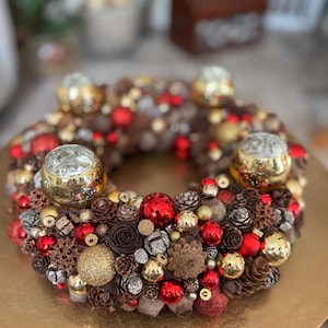 Könnte beinhalten: Ein festlicher Weihnachtskranz mit kreisförmigem Design, verziert mit roten und goldenen Ornamenten, Tannenzapfen und vier Glaskerzenhaltern. Der Kranz steht auf einer goldenen Oberfläche und schafft eine warme und einladende Weihnachtsdekoration.