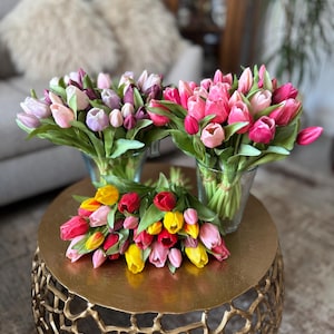 Könnte beinhalten: Drei Sträuße aus künstlichen Tulpen. Ein Strauß besteht aus hellvioletten und rosafarbenen Blumen in einer Glasvase. Ein weiterer Strauß enthält leuchtend rosa Tulpen in einer Glasvase. Ein dritter Strauß aus gelben, roten und rosa Tulpen liegt flach.