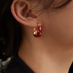 May include: Gold hoop earrings with red enamel. The earrings are a simple design with a smooth, rounded shape.