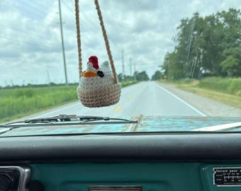 May include: A crocheted chicken car charm hanging from a rearview mirror. The chicken has a red comb, orange beak, and white and beige body. The car's dashboard is teal, and the road ahead is visible through the windshield.