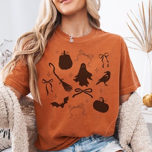 May include: A burnt orange short-sleeve t-shirt featuring a dark brown graphic print of various Halloween-themed icons including pumpkins, a ghost, a broomstick, a raven, a bat, and several decorative bows interspersed with small star shapes.