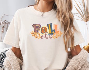 May include: Ivory-colored t-shirt with the text "Fall Vibes" in a colorful, decorative font. The design includes autumn leaves. The model is wearing denim shorts and a beige cardigan.