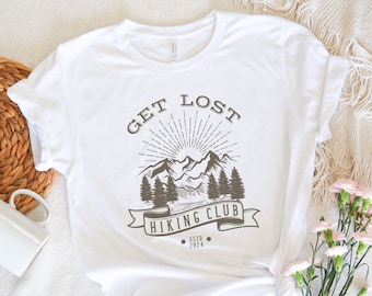 May include: White t-shirt with a brown graphic design that says "Get Lost Hiking Club Est. 2024" with a mountain range and sun illustration.