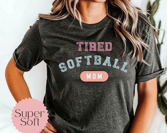 May include: A dark grey t-shirt with the text "TIRED SOFTBALL MOM" in pink and blue lettering. The text has a distressed, vintage look. The shirt is super soft.