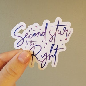May include: A white sticker with the text "Second Star to the Right" in a cursive font, with a gradient of purple and blue. The sticker has a glossy finish and is held by a hand.