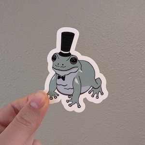 May include: A green frog wearing a black top hat and bow tie. The frog is sitting on a white background.