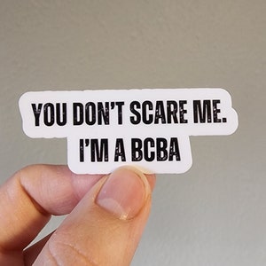 May include: A white sticker with black text that reads "YOU DON'T SCARE ME. I'M A BCBA." The sticker is held by a person's fingers against a light gray background. The sticker has a unique shape.