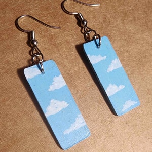 May include: A pair of blue and white painted wooden earrings with silver hooks. The earrings are shaped like rectangles and feature a sky blue background with white clouds painted on them.
