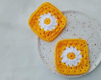 May include: Two handmade crochet coasters. Each coaster has a daisy design with a white flower centre and yellow petals. The square coasters sit on a speckled white plate.