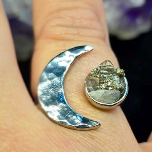 May include: A silver ring featuring a crescent moon design and a round setting with a cluster of crystals. The ring has a hammered texture on the moon and a unique, eye-catching style. The ring is a piece of jewelry.