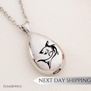 Op de afbeelding: Een zilveren, traanvormige ketting met een zwarte haai-silhouetontwerp. De hanger heeft een kleine opening aan de onderkant.