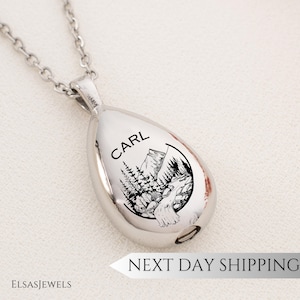 Peut inclure: Un collier pendentif argenté en forme de goutte d'eau. Le pendentif présente un paysage de montagne et de forêt en noir et blanc, avec le nom "CARL" au-dessus. Le collier a une chaîne fine et une petite ouverture en bas.