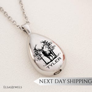 Op de afbeelding: Een zilverkleurige ketting met een druppelvormige hanger. De hanger heeft een zwarte illustratie van een hert met bomen en de naam "TYLER". De ketting heeft een ketting en een sluiting.