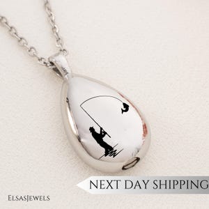 May include: A silver-colored teardrop-shaped pendant necklace with a chain. The pendant features a black silhouette of a person fishing, with a fish jumping. The text "NEXT DAY SHIPPING" is visible.