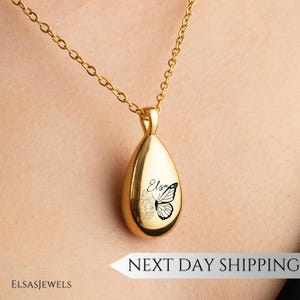 Op de afbeelding: Een goudkleurige, druppelvormige hangerketting. De hanger heeft een zwart vlinderontwerp met de naam "Elsa" en bloemaccenten. De ketting hangt aan een fijne gouden ketting.