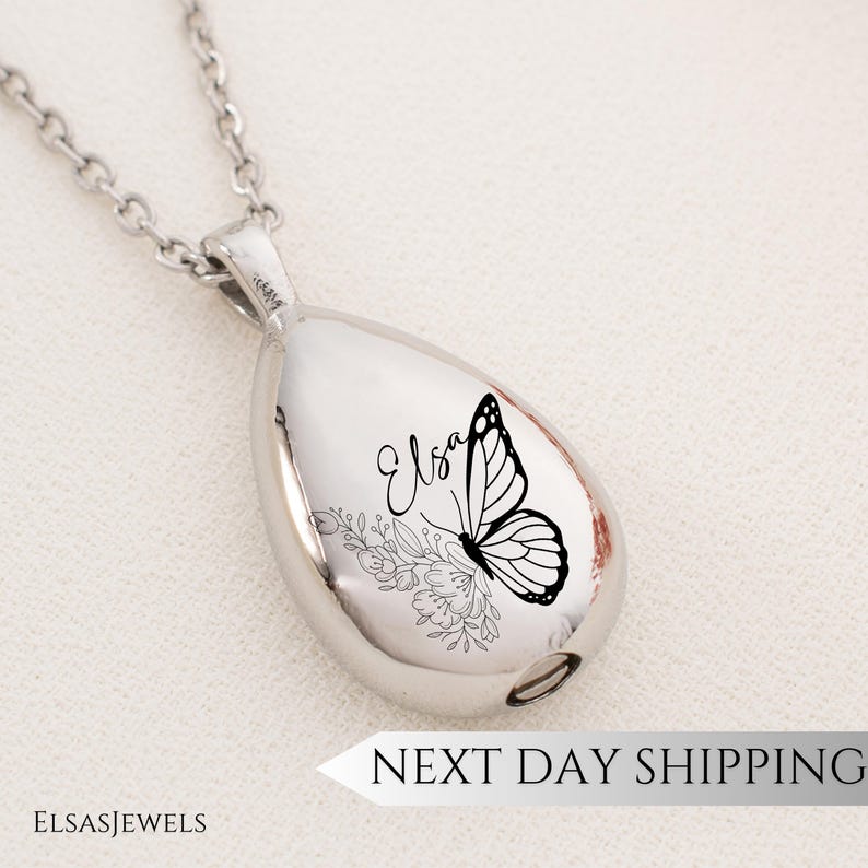 Op de afbeelding: Een zilverkleurige ketting met een druppelvormige hanger. De hanger heeft een zwart vlinder- en bloemmotief met de naam "Elsa". De ketting heeft een fijne ketting. De tekst "NEXT DAY SHIPPING" is zichtbaar.