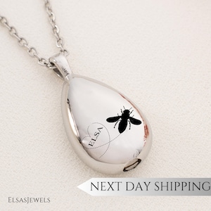May include: A silver-colored teardrop-shaped pendant necklace. The pendant features a black bee design with the name "ELSA" inside a heart. The necklace has a chain and a clasp. The brand name "ELSASJEWELS" is at the bottom.