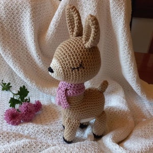 May include: A crocheted deer toy with brown fur, white face details, and a pink scarf. The deer is standing on a white crocheted blanket.