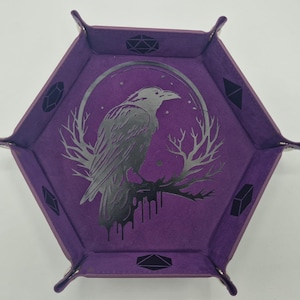 Può includere: Vassoio per dadi esagonale viola con un'illustrazione di corvo argentato. Il corvo è appollaiato su un ramo all'interno di una cornice circolare. Il vassoio ha angoli rinforzati e forme geometriche sui lati.