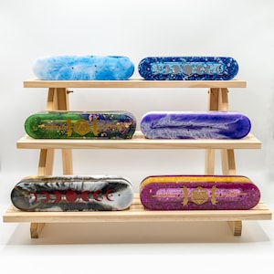 Puede incluir: Seis cajas ovaladas de resina de colores, expuestas en una estantería de madera de varios niveles. Las cajas presentan varios diseños, incluyendo remolinos azules, patrones celestiales y detalles dorados. Cada caja es única, mostrando una variedad de colores y texturas.