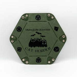 Puede incluir: Una bandeja verde hexagonal para dados con un diseño de silueta de gato negro y el texto "DUNGEON MASTER CAT HERDER". La bandeja tiene cierres de presión plateados y símbolos de dados en los bordes. Un divertido accesorio para juegos de mesa.