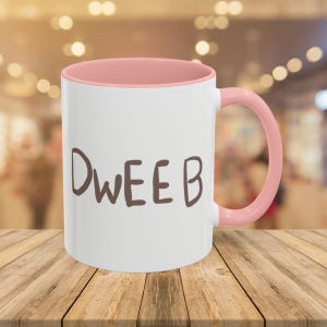 May include: A white ceramic mug with a pink rim and handle. The mug has the word "DWEEB" written on it in brown lettering.