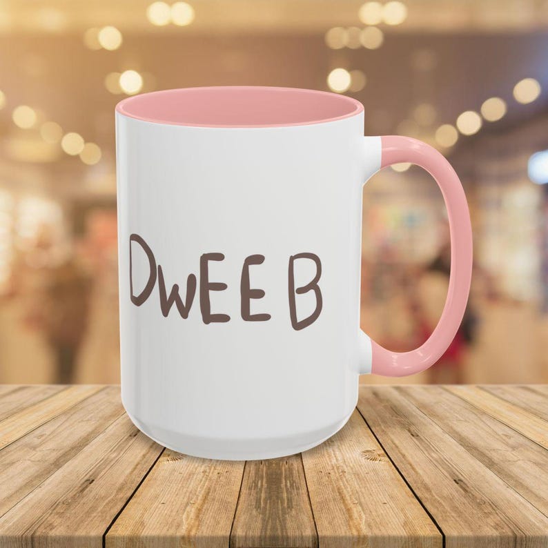 May include: White ceramic coffee mug with a pink rim and handle. The mug has the word "DWEEB" written in brown on the front.