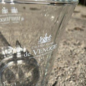 May include: A clear glass champagne bucket with the words "CHAMPAGNE de VENOGE Depuis 1837" printed in white. The bucket has a trapezoidal shape and a wide opening. The logo is a crest with two lions.