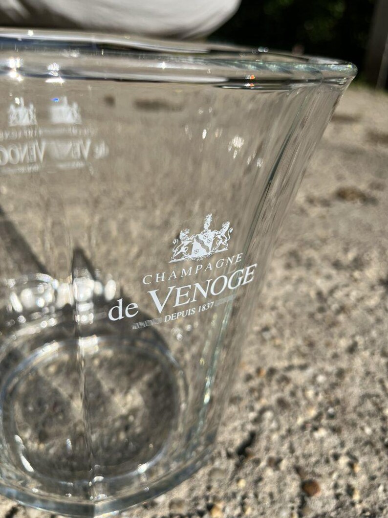 May include: A clear glass champagne bucket with the words "CHAMPAGNE de VENOGE Depuis 1837" printed in white. The bucket has a trapezoidal shape and a wide opening. The logo is a crest with two lions.