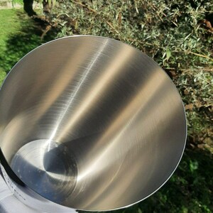 Pode incluir: Uma tigela de mistura de a&ccedil;o inoxid&aacute;vel com interior polido. A tigela &eacute; segurada por uma m&atilde;o enluvada, com um fundo de folhagem verde e luz solar. O interior da tigela reflete a luz, criando um brilho met&aacute;lico brilhante.