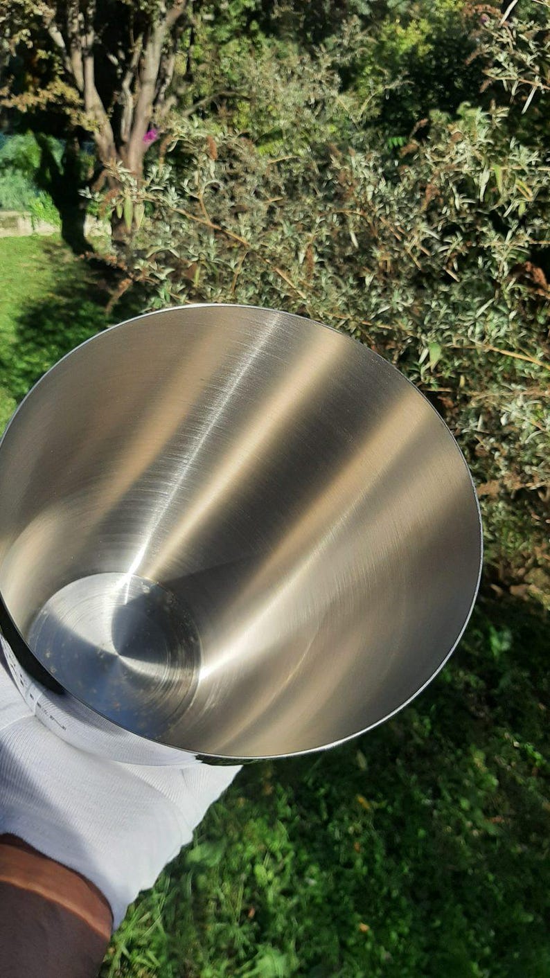 Pode incluir: Uma tigela de mistura de a&ccedil;o inoxid&aacute;vel com interior polido. A tigela &eacute; segurada por uma m&atilde;o enluvada, com um fundo de folhagem verde e luz solar. O interior da tigela reflete a luz, criando um brilho met&aacute;lico brilhante.
