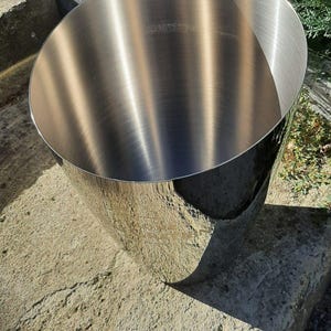 Pode incluir: Um balde de gelo de metal inoxid&aacute;vel com acabamento polido. O balde tem uma parte superior larga e aberta e uma forma cil&iacute;ndrica. A superf&iacute;cie reflete a luz solar, criando destaques e sombras. O balde est&aacute; sobre uma superf&iacute;cie de pedra.