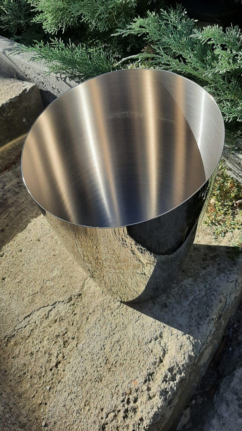 Pode incluir: Um balde de gelo de metal inoxid&aacute;vel com acabamento polido. O balde tem uma parte superior larga e aberta e uma forma cil&iacute;ndrica. A superf&iacute;cie reflete a luz solar, criando destaques e sombras. O balde est&aacute; sobre uma superf&iacute;cie de pedra.