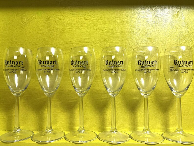 May include: Six clear champagne flutes with the "Ruinart Champagne" logo printed on the front. The glasses are arranged in a row against a yellow background. The text "Champagne" is visible on the glasses.