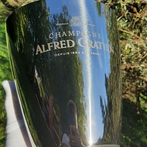 Pode incluir: Balde de champanhe prateado e brilhante com as palavras "CHAMPAGNE ALFRED GRATIN" e "DEPUIS 1864 A. CERNAY". O balde reflete o ambiente. Um acess&oacute;rio elegante para servir champanhe.