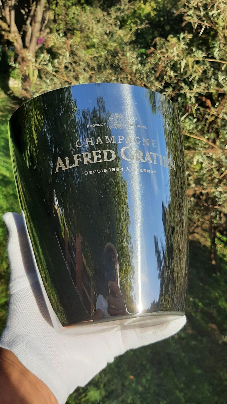 Pode incluir: Balde de champanhe prateado e brilhante com as palavras "CHAMPAGNE ALFRED GRATIN" e "DEPUIS 1864 A. CERNAY". O balde reflete o ambiente. Um acess&oacute;rio elegante para servir champanhe.