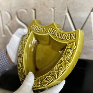 K&ouml;nnte beinhalten: Eine schildf&ouml;rmige, olivgr&uuml;ne Keramikschale mit den erhabenen Worten "MO&Euml;T & CHANDON". Die Schale hat ein Trauben- und Blattmuster an den R&auml;ndern. Die Schale wird von behandschuhten H&auml;nden gehalten.