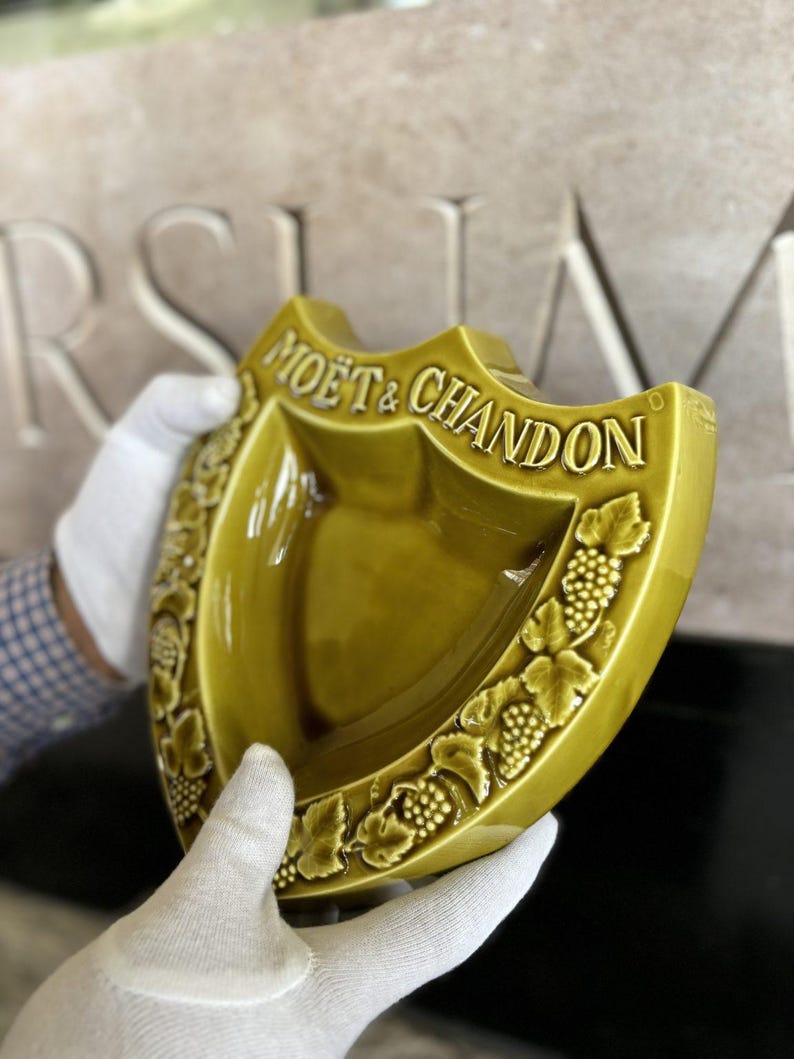 K&ouml;nnte beinhalten: Eine schildf&ouml;rmige, olivgr&uuml;ne Keramikschale mit den erhabenen Worten "MO&Euml;T & CHANDON". Die Schale hat ein Trauben- und Blattmuster an den R&auml;ndern. Die Schale wird von behandschuhten H&auml;nden gehalten.