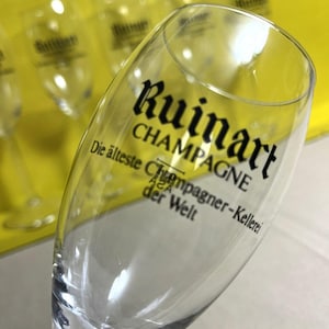 May include: A clear champagne flute with the word "Ruinart" printed in black. The text "CHAMPAGNE" is below the brand name. Additional text in German is also visible. Several other flutes are in the background.