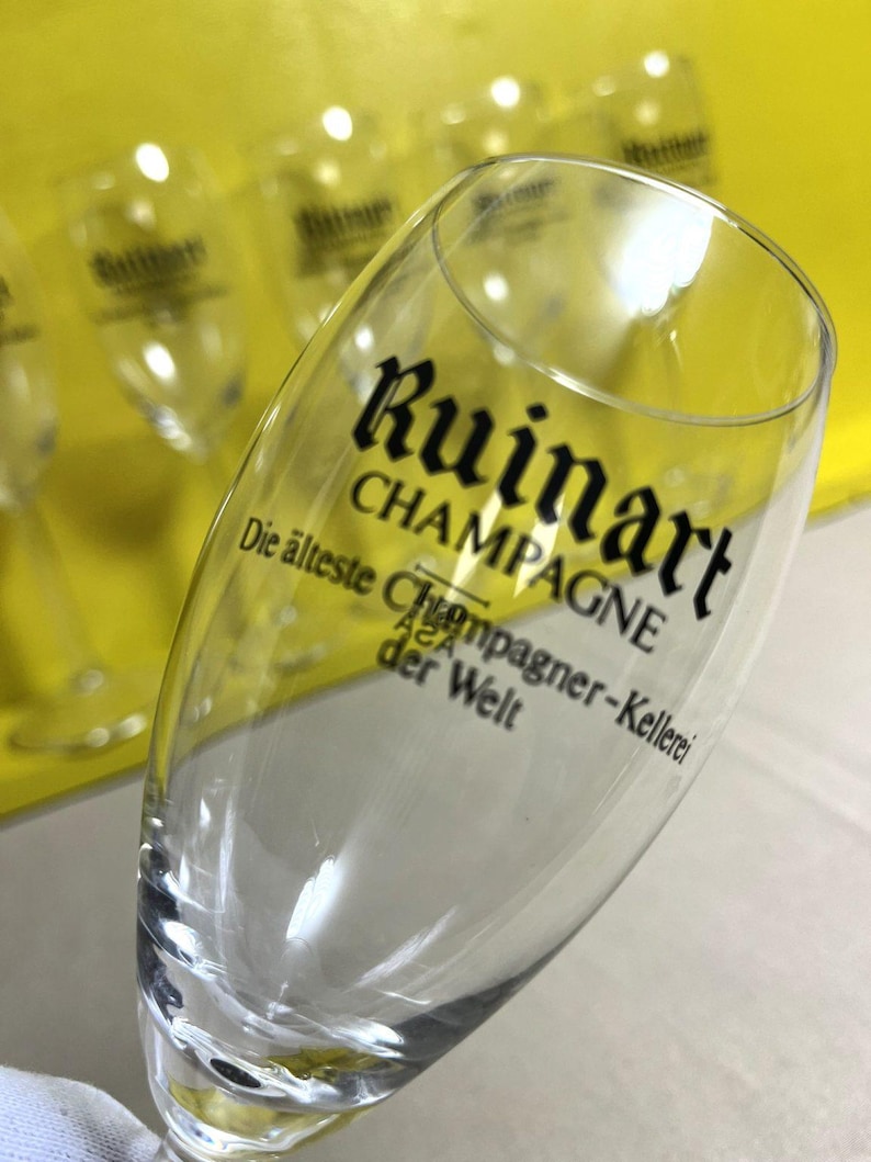 May include: A clear champagne flute with the word "Ruinart" printed in black. The text "CHAMPAGNE" is below the brand name. Additional text in German is also visible. Several other flutes are in the background.