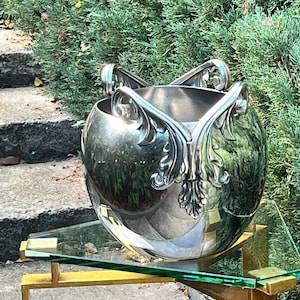 Puede incluir: Un cuenco decorativo plateado, de forma esf&eacute;rica, con adornos florales estilizados. Se apoya sobre una balda de cristal sostenida por un marco met&aacute;lico dorado. El fondo muestra follaje verde y escalones de piedra.