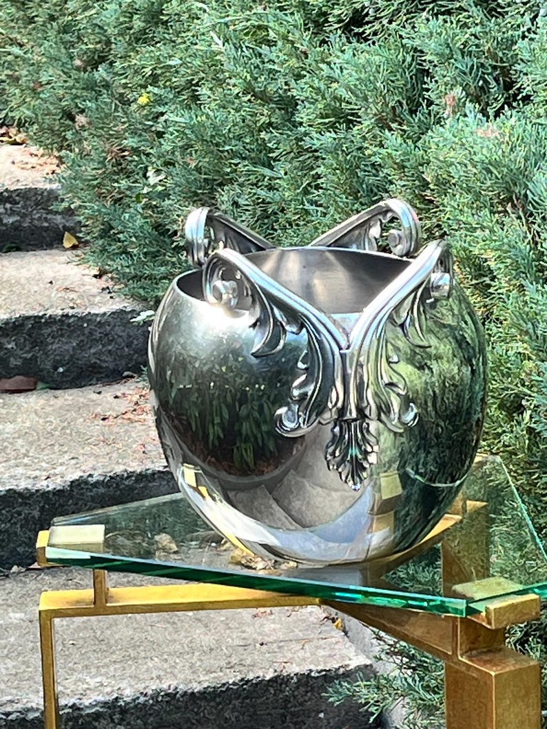 Puede incluir: Un cuenco decorativo plateado, de forma esf&eacute;rica, con adornos florales estilizados. Se apoya sobre una balda de cristal sostenida por un marco met&aacute;lico dorado. El fondo muestra follaje verde y escalones de piedra.