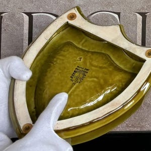 K&ouml;nnte beinhalten: Eine schildf&ouml;rmige, olivgr&uuml;ne Keramikschale mit cremefarbenem Rand und messingfarbenen Nieten. Die Schale hat eine gl&auml;nzende Oberfl&auml;che und ein strukturiertes Inneres. Der Boden der Schale hat einen schwarzen Stempel mit Text.