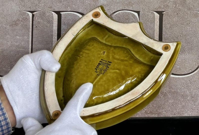 K&ouml;nnte beinhalten: Eine schildf&ouml;rmige, olivgr&uuml;ne Keramikschale mit cremefarbenem Rand und messingfarbenen Nieten. Die Schale hat eine gl&auml;nzende Oberfl&auml;che und ein strukturiertes Inneres. Der Boden der Schale hat einen schwarzen Stempel mit Text.