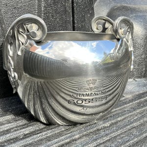 Puede incluir: Una cubitera de champ&aacute;n plateada con adornos de volutas y las palabras "CHAMPAGNE GOSSET" en relieve en la parte delantera. La cubitera tiene una superficie reflectante y asas decorativas.