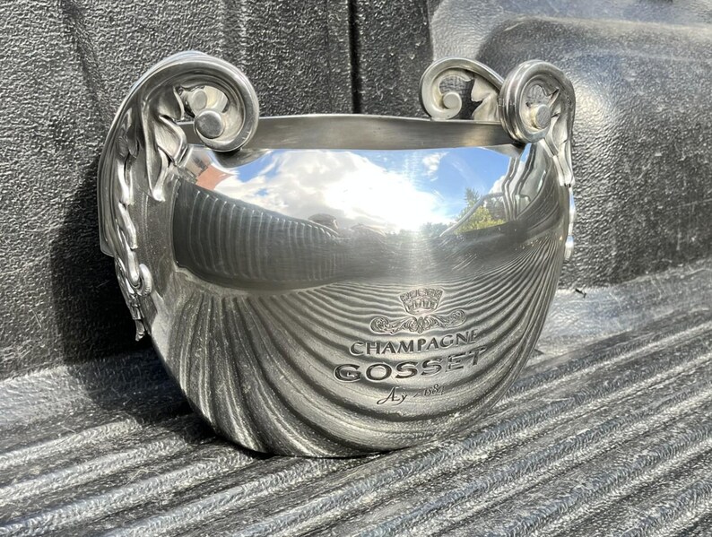 Puede incluir: Una cubitera de champ&aacute;n plateada con adornos de volutas y las palabras "CHAMPAGNE GOSSET" en relieve en la parte delantera. La cubitera tiene una superficie reflectante y asas decorativas.