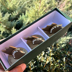 May include: A set of four decorative, brown and gold-colored coasters with a leaf-like design, each embellished with small, clear rhinestones. The coasters are presented in a green box with white dividers, ideal for home decor or gifting.
