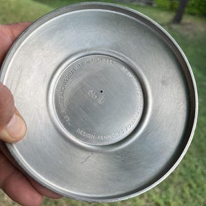 Puede incluir: Un objeto circular de metal cepillado con un centro ligeramente c&oacute;ncavo. Los textos "LE RAFRAICHISSEUR DE MEMOIRES" y "DESIGN PENNOR'S POUR NICOLAS FEUILLATTE" est&aacute;n grabados alrededor del centro. El objeto tiene un acabado plateado mate y parece ser una pieza de servicio.