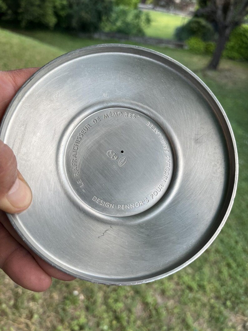 Puede incluir: Un objeto circular de metal cepillado con un centro ligeramente c&oacute;ncavo. Los textos "LE RAFRAICHISSEUR DE MEMOIRES" y "DESIGN PENNOR'S POUR NICOLAS FEUILLATTE" est&aacute;n grabados alrededor del centro. El objeto tiene un acabado plateado mate y parece ser una pieza de servicio.