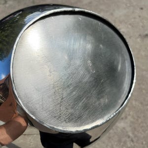 Puede incluir: Un recipiente de metal plateado brillante con forma redondeada. La parte inferior del recipiente tiene un dise&ntilde;o circular con ara&ntilde;azos visibles. El recipiente refleja el entorno, incluidos &aacute;rboles y cielo.